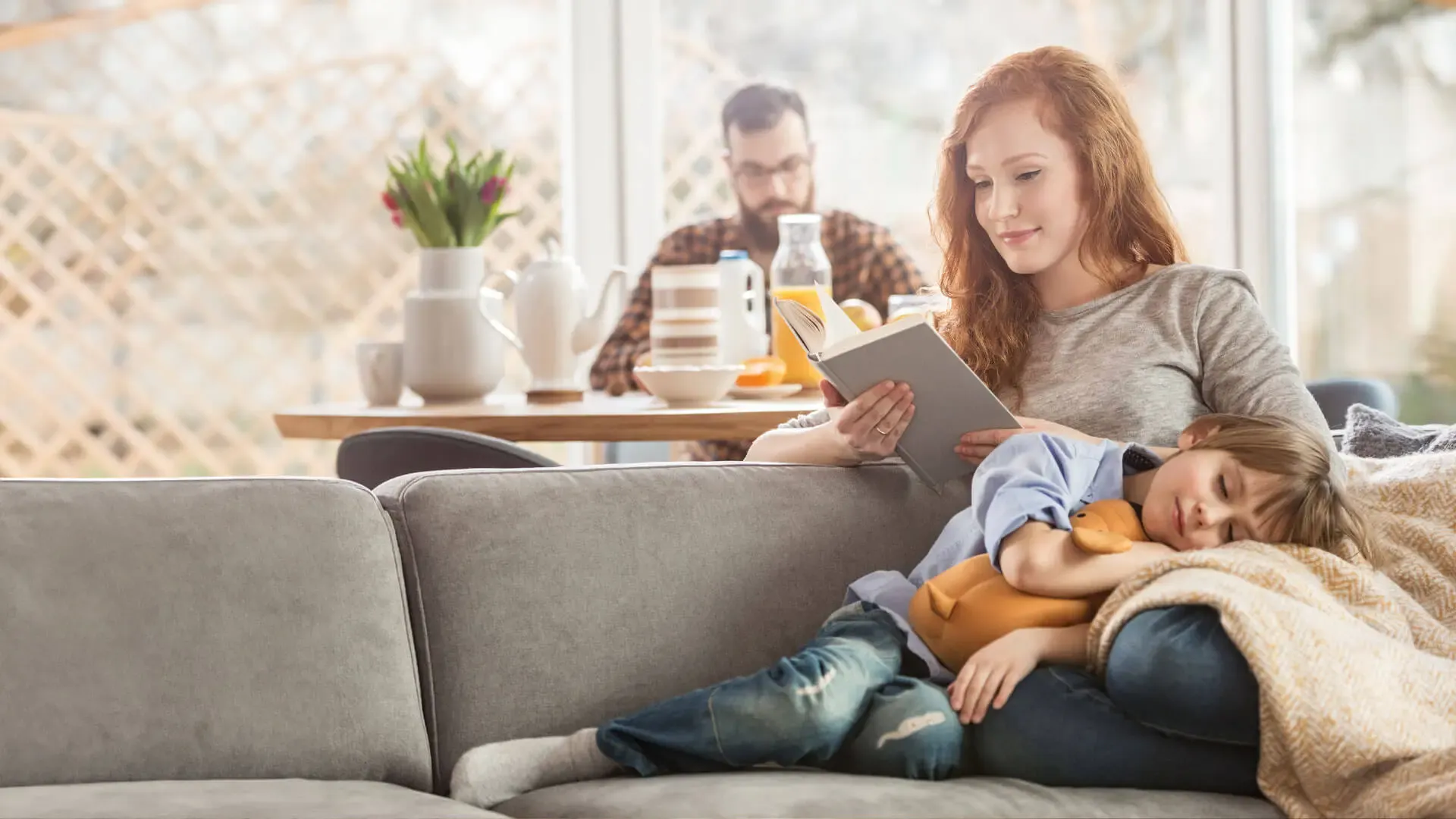 Frau sitzt mit Kind auf der Couch und schaut aufs Tablet, Mann im Hintergrund