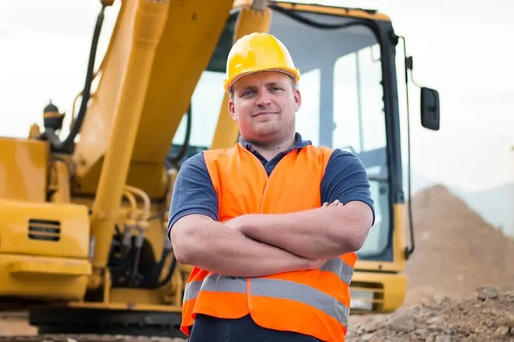 Mann mit PSA vor Bagger auf Tiefbaustelle